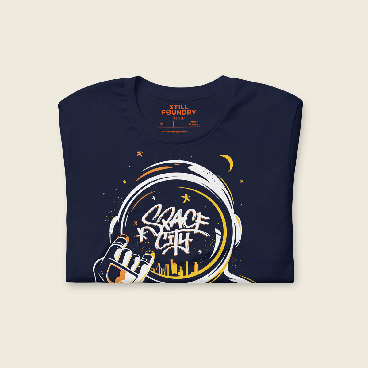 Folded Navy t-shirt featuring an illustrated astronaut holding up the H-Town hand sign, with the word “Space City” written in graffiti-style lettering inside the astronaut’s helmet and a yellow Houston skyline reflected below, surrounded by stars and a crescent moon.