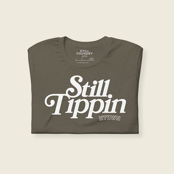 Army green t-shirt folded with the phrase "Still Tippin HTOWN" printed in bold white lettering on the front. The text features a stylish, retro font, with "Still Tippin" in large letters and "HTOWN" in smaller block letters underneath. 