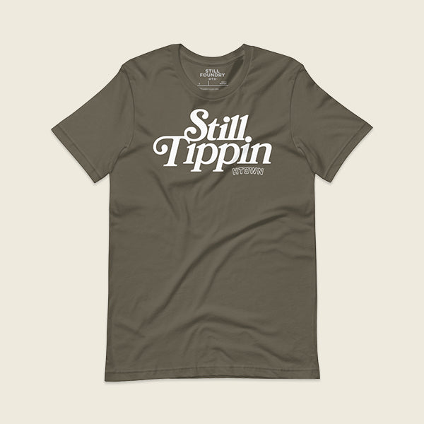 Army green t-shirt with the phrase "Still Tippin HTOWN" printed in bold white lettering on the front. The text features a stylish, retro font, with "Still Tippin" in large letters and "HTOWN" in smaller block letters underneath. 