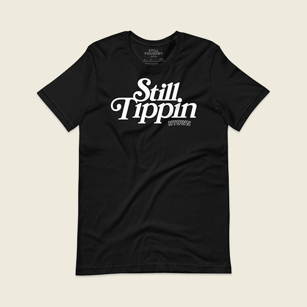 Black t-shirt with the phrase "Still Tippin HTOWN" printed in bold white lettering on the front. The text features a stylish, retro font, with "Still Tippin" in large letters and "HTOWN" in smaller block letters underneath. 