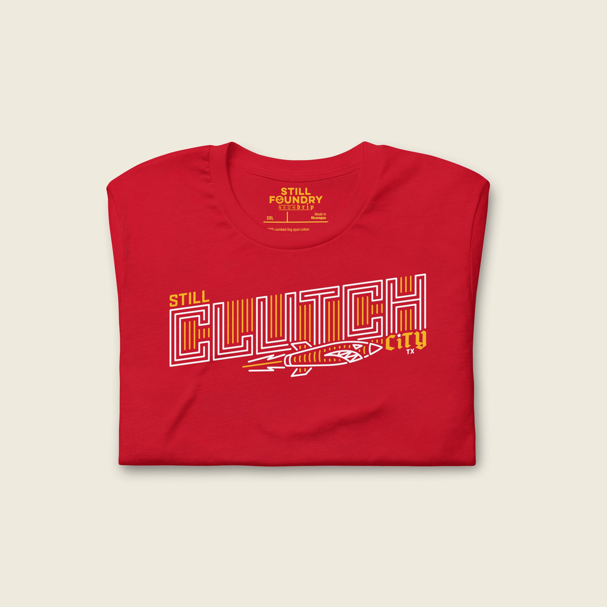Folded red t-shirt featuring a bold, outlined graphic that reads “Still Clutch City” in stylized white and yellow lettering, with a dynamic rocket illustration underneath.