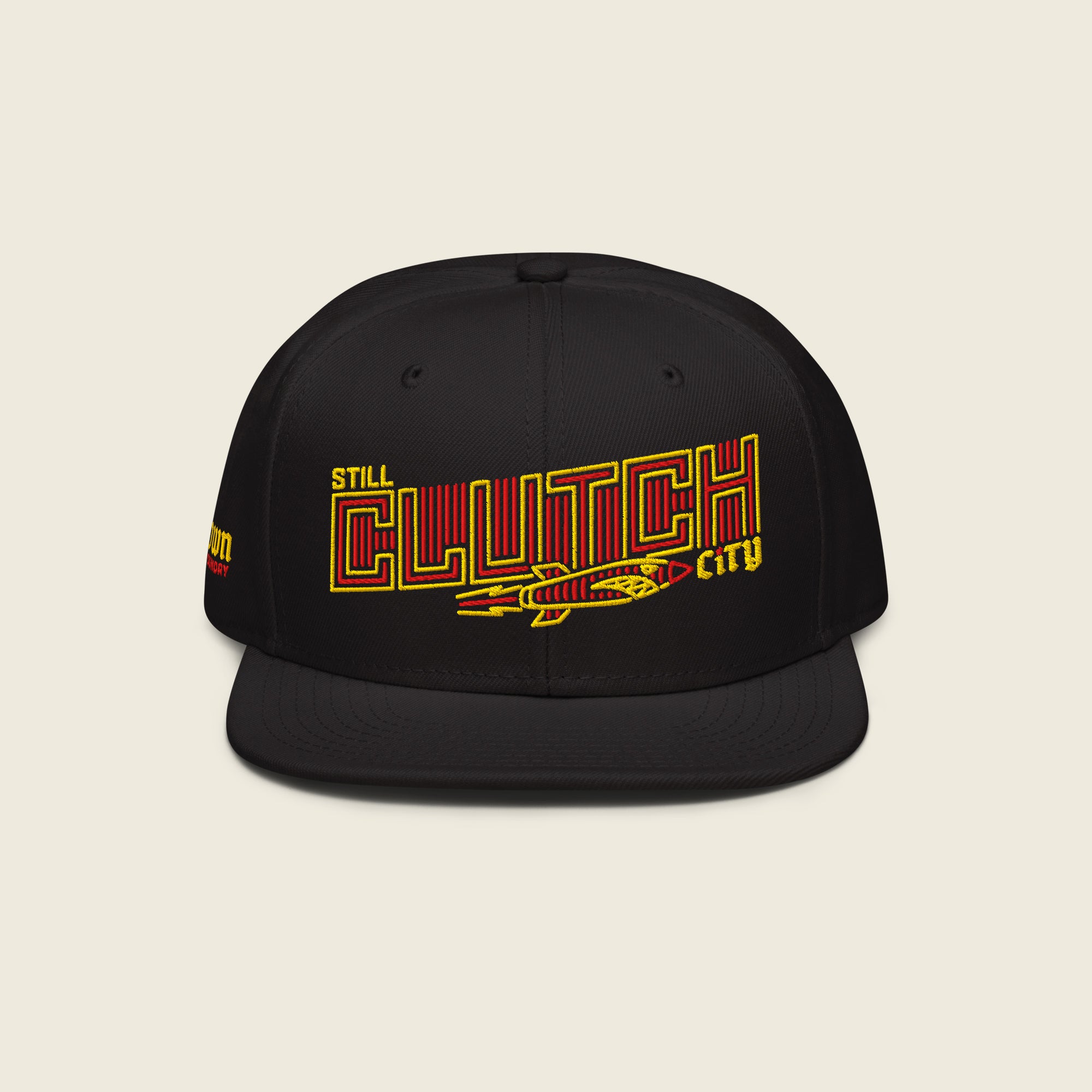 Black structured snapback hat with flat bill; front embroidery reads “STILL” (small yellow) above a large outlined “CLUTCH” filled with red vertical stripes, with a vintage rocket beneath and a small yellow “city” at the right.