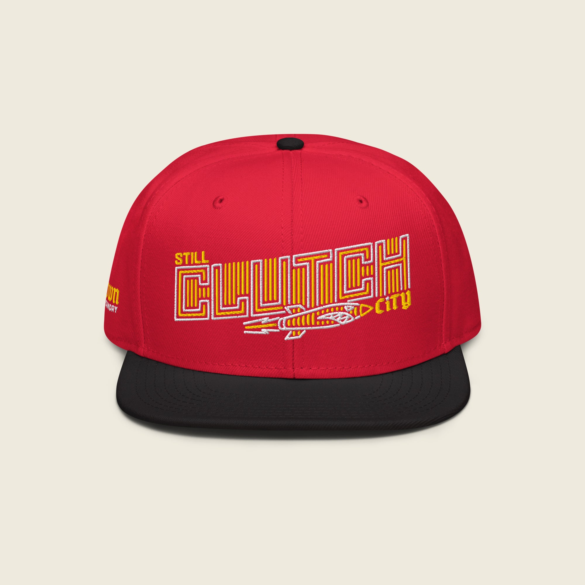 Red structured snapback hat with a black flat bill; front embroidery reads “STILL” (small yellow) above a large outlined “CLUTCH” filled with yellow vertical stripes, with a vintage rocket beneath and a small yellow “city” at the right.