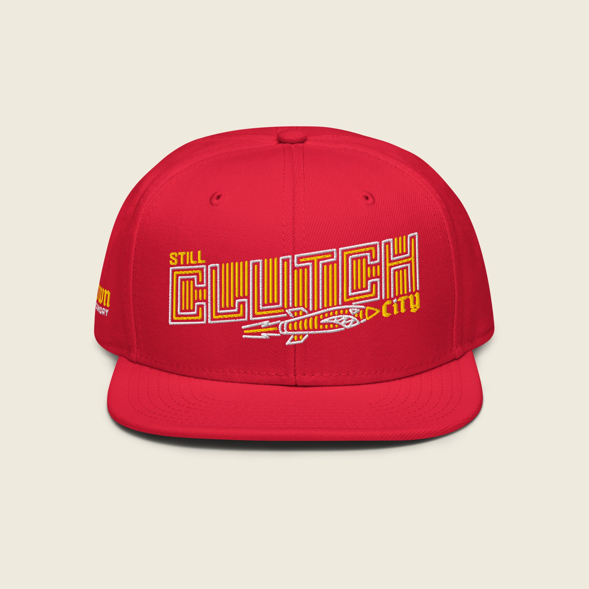 Red structured snapback hat with flat bill; front embroidery reads “STILL” (small yellow) above a large outlined “CLUTCH” filled with yellow vertical stripes, with a vintage rocket beneath and a small yellow “city” at the right.