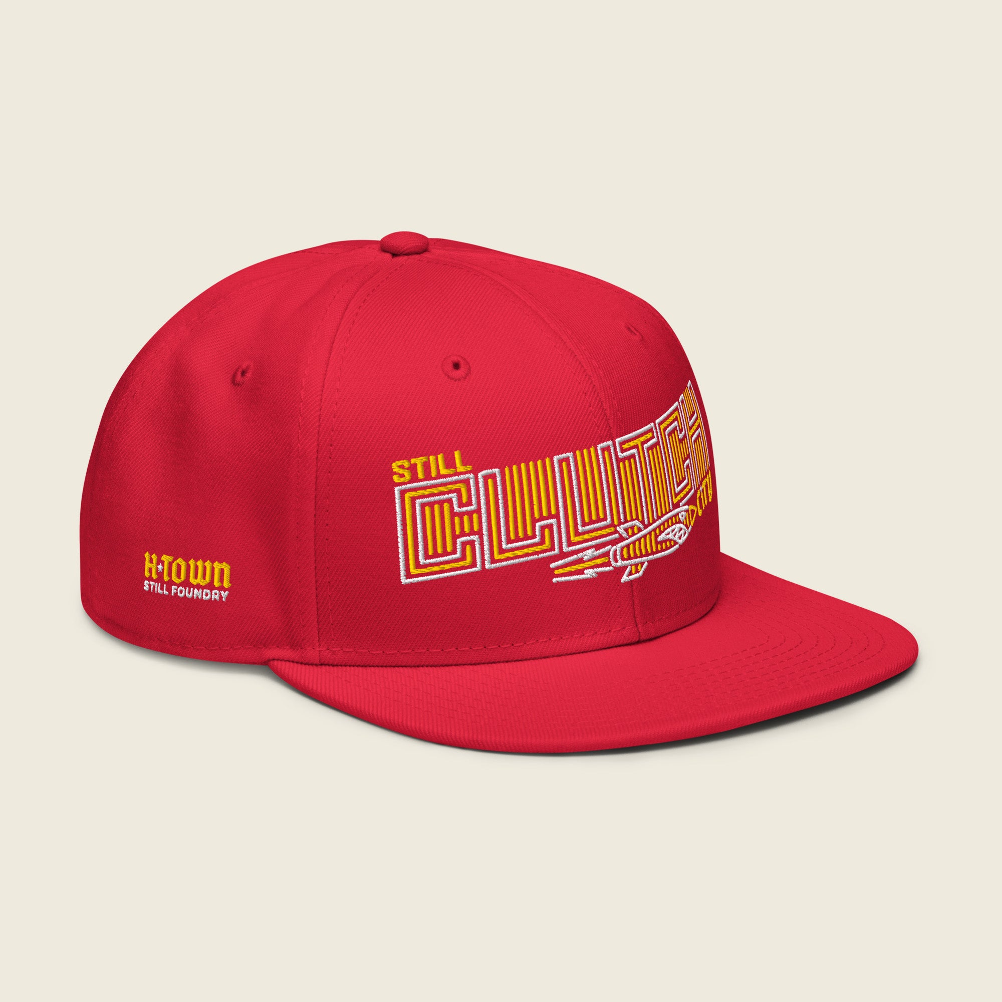 Red structured snapback hat with flat bill; front embroidery reads “STILL” (small yellow) above a large outlined “CLUTCH” filled with yellow vertical stripes, with a vintage rocket beneath and a small yellow “city” at the right.
