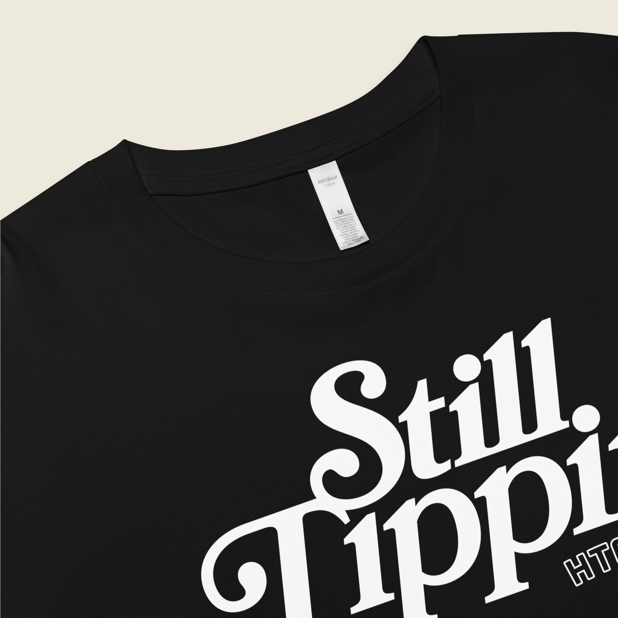 Black crop top with the phrase "Still Tippin HTOWN" printed in bold white lettering on the front. The text features a stylish, retro font, with "Still Tippin" in large letters and "HTOWN" in smaller block letters underneath. 