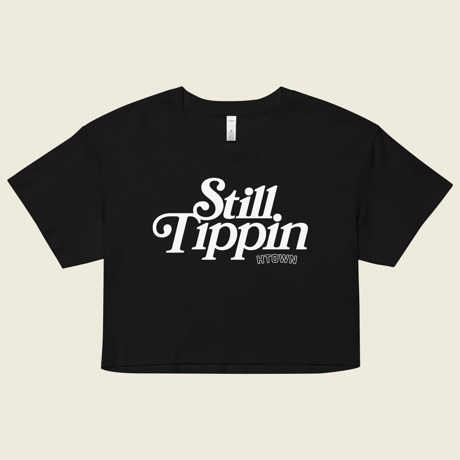 Black crop top with the phrase "Still Tippin HTOWN" printed in bold white lettering on the front. The text features a stylish, retro font, with "Still Tippin" in large letters and "HTOWN" in smaller block letters underneath. 