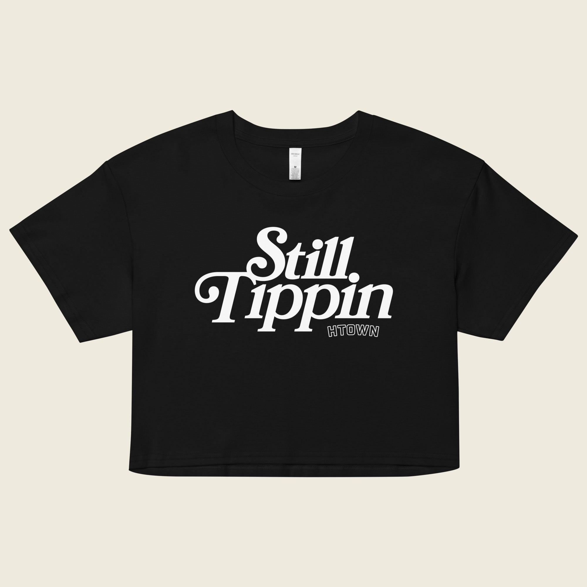 Black crop top with the phrase "Still Tippin HTOWN" printed in bold white lettering on the front. The text features a stylish, retro font, with "Still Tippin" in large letters and "HTOWN" in smaller block letters underneath. 