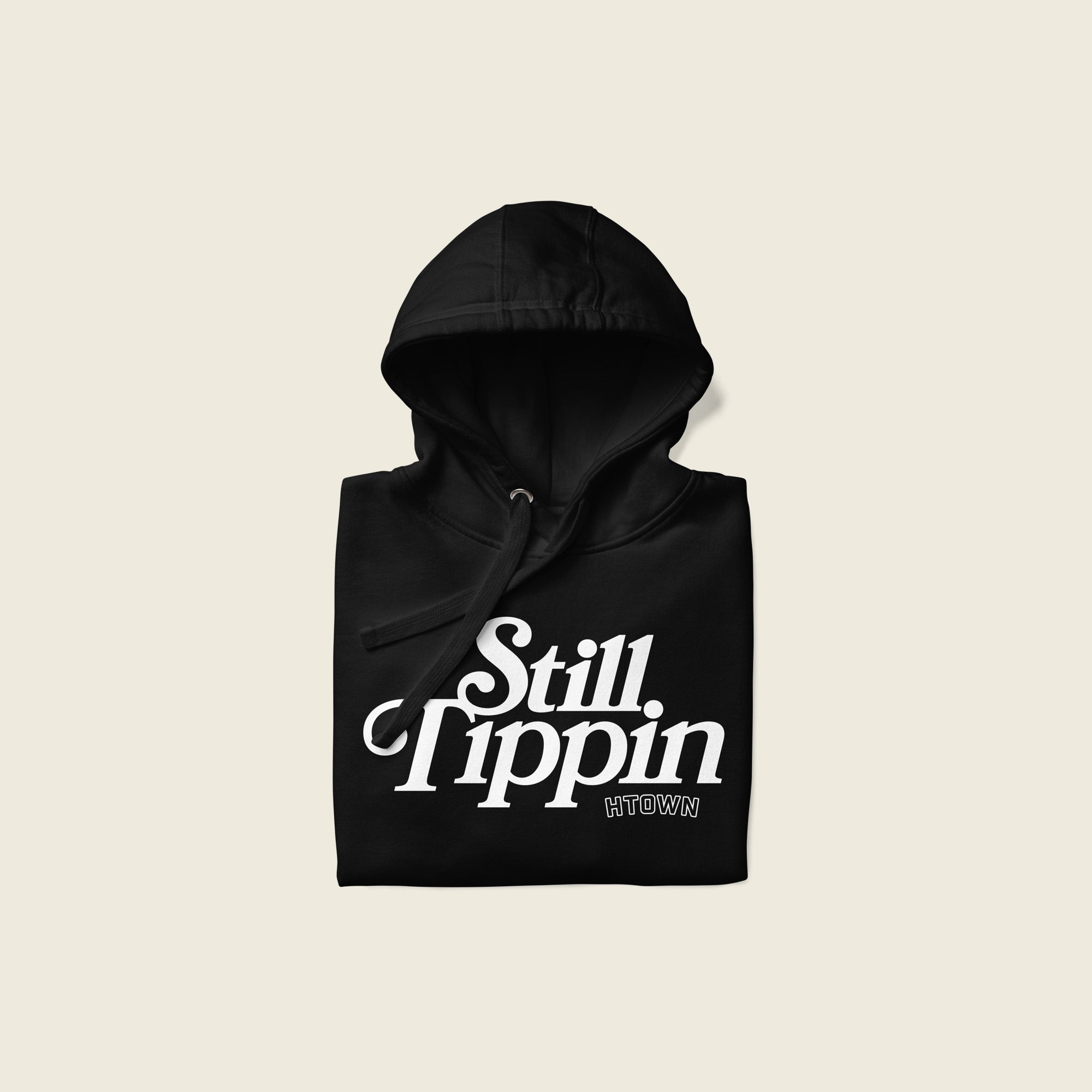 Black hoodie folded with the phrase "Still Tippin HTOWN" printed in bold white lettering on the front. The text features a stylish, retro font, with "Still Tippin" in large letters and "HTOWN" in smaller block letters underneath. 